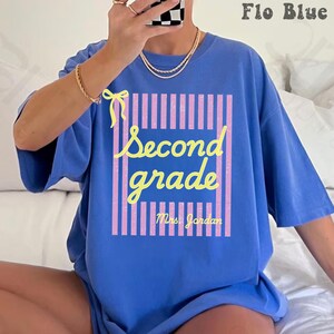 Puede incluir: Una camiseta azul con un gráfico de rayas verticales rosas, un lazo amarillo y el texto amarillo Second grade Mrs. Jordan. El diseño está centrado en el pecho de la camiseta. La prenda tiene mangas cortas y un cuello redondo.