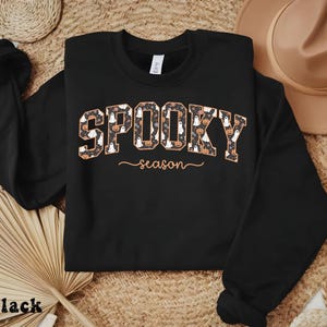 Black sweatshirt with spooky season design featuring ghosts, pumpkins, and flowers, ideal for fall outfits or Halloween gift.