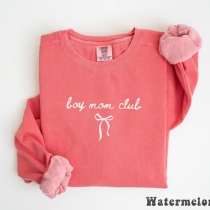 K&ouml;nnte beinhalten: Ein korallenfarbenes Sweatshirt mit dem wei&szlig;en Schriftzug "boy mom club" und einer Schleifen-Grafik. Das lang&auml;rmlige Oberteil hat einen Rundhalsausschnitt. Unten steht das Wort "Watermelon".