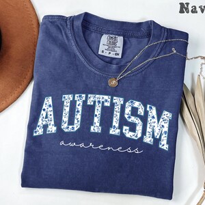 May include: Navy blue t-shirt with the word "AUTISM" in white block letters filled with a blue floral pattern. The word "awareness" is written in cursive below. A gold necklace rests on the shirt.