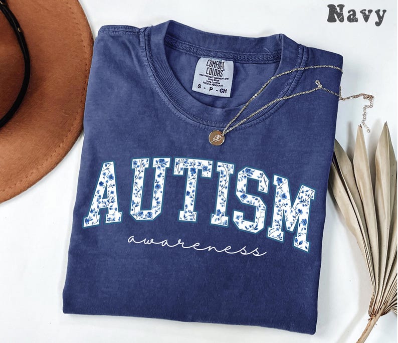 May include: Navy blue t-shirt with the word "AUTISM" in white block letters filled with a blue floral pattern. The word "awareness" is written in cursive below. A gold necklace rests on the shirt.