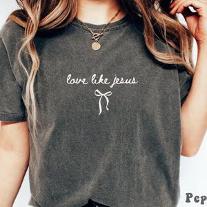 May include: Dark gray t-shirt with the phrase "love like jesus" in white script and a bow graphic. The shirt has short sleeves and a crew neck. The word "Pepper" is printed in the bottom right corner.