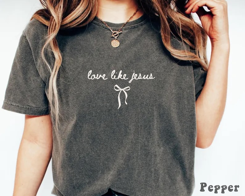 May include: Dark gray t-shirt with the phrase "love like jesus" in white script and a bow graphic. The shirt has short sleeves and a crew neck. The word "Pepper" is printed in the bottom right corner.
