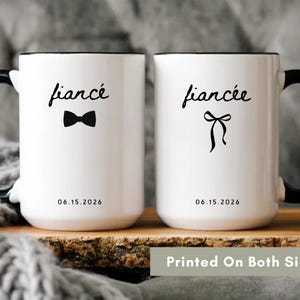 Puede incluir: Dos tazas de cerámica blancas con asas y bordes negros. Una taza tiene la palabra "fiancé" con una pajarita, la otra "fiancée" con un lazo. Ambas tazas tienen la fecha 06.15.2026.