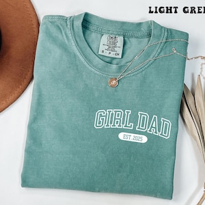 May include: Light green t-shirt with the text "GIRL DAD EST. 2025" in white block letters. A gold necklace with a pendant rests on the shirt. The shirt is folded and placed on a white surface.