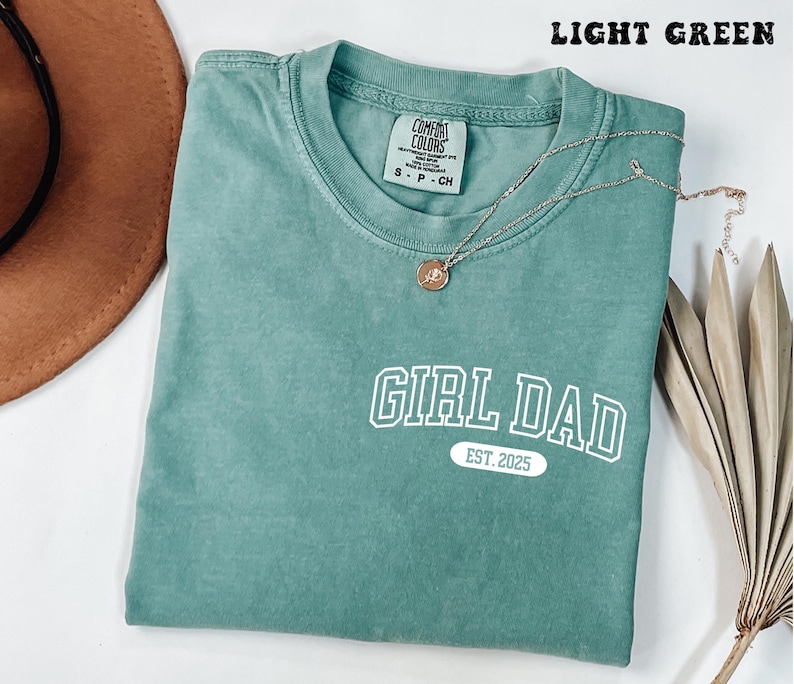 May include: Light green t-shirt with the text "GIRL DAD EST. 2025" in white block letters. A gold necklace with a pendant rests on the shirt. The shirt is folded and placed on a white surface.