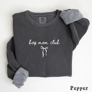 K&ouml;nnte beinhalten: Ein dunkelgraues Sweatshirt mit dem wei&szlig;en Schriftzug "boy mom club" und einer Schleifengrafik. Die &Auml;rmel sind mit einem helleren, flauschigen Material versehen. Das Wort "Pepper" steht unten.