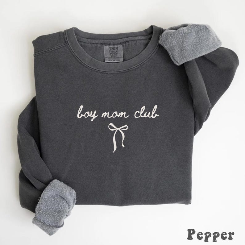 K&ouml;nnte beinhalten: Ein dunkelgraues Sweatshirt mit dem wei&szlig;en Schriftzug "boy mom club" und einer Schleifengrafik. Die &Auml;rmel sind mit einem helleren, flauschigen Material versehen. Das Wort "Pepper" steht unten.