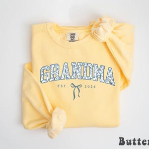 May include: A light yellow sweatshirt with the word "GRANDMA" in a blue floral design. Below the word is "EST. 2026" and a bow graphic. The word "Butter" is at the bottom right.