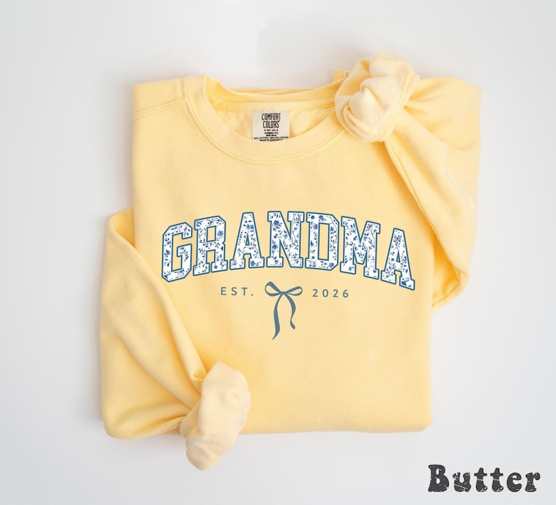 May include: A light yellow sweatshirt with the word "GRANDMA" in a blue floral design. Below the word is "EST. 2026" and a bow graphic. The word "Butter" is at the bottom right.