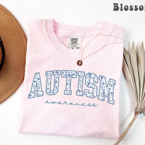 May include: A light pink t-shirt with the word "AUTISM" in blue floral print, and the word "awareness" in a script font. A gold necklace with a pendant is on the shirt. The word "Blossom" is at the top right.