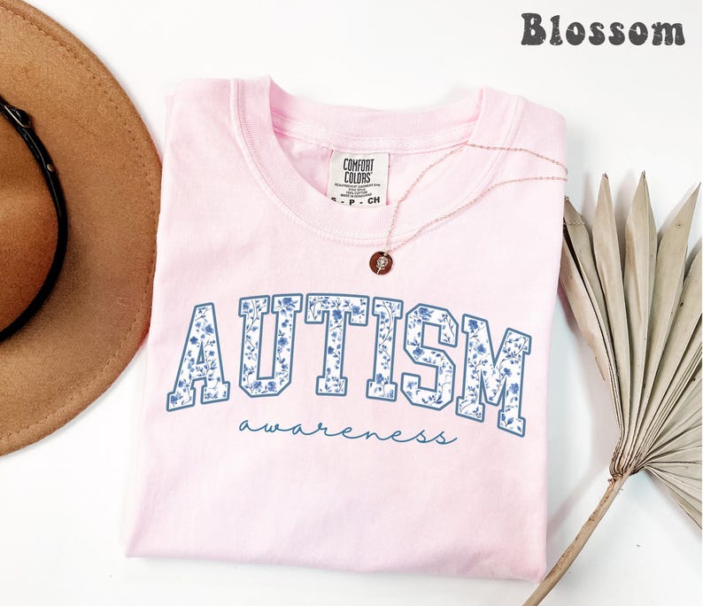 May include: A light pink t-shirt with the word "AUTISM" in blue floral print, and the word "awareness" in a script font. A gold necklace with a pendant is on the shirt. The word "Blossom" is at the top right.