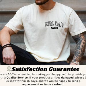 May include: Cream-colored t-shirt with the text "GIRL DAD EST. 2025" in black, featuring a classic collegiate font. The shirt is short-sleeved and has a relaxed fit. The image shows the shirt on a person sitting on steps.