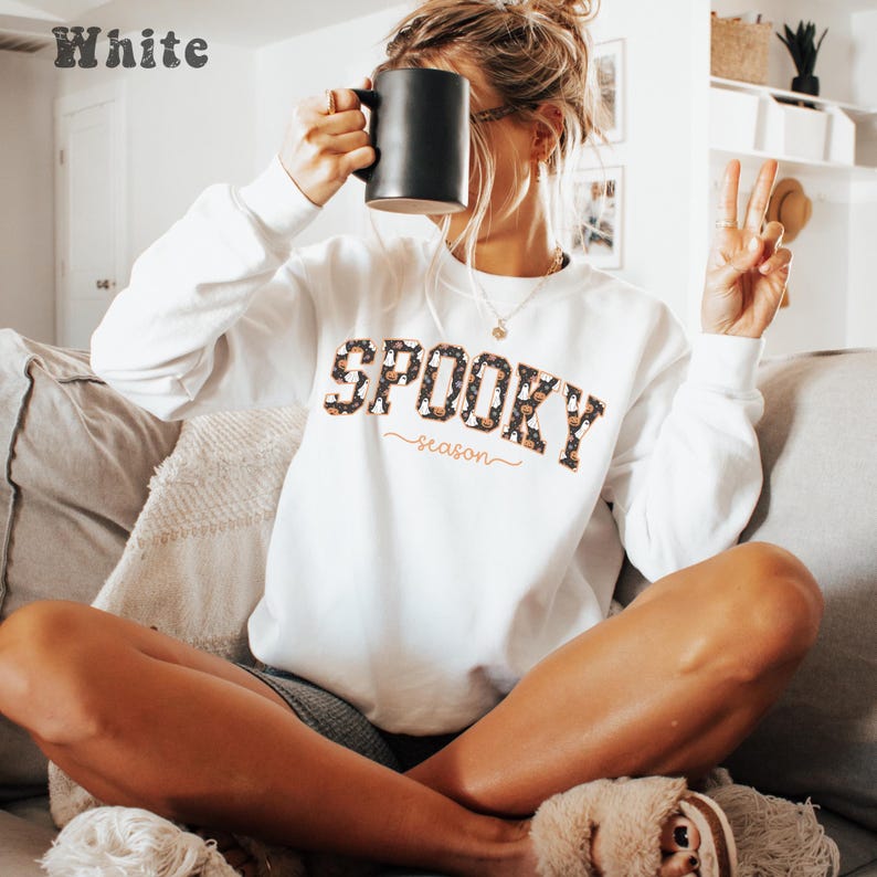 May include: White sweatshirt with the text "SPOOKY season" in a leopard print design. The sweatshirt is long-sleeved and has a crew neck. A black mug is held up to the face. The word "White" is in the top left corner.