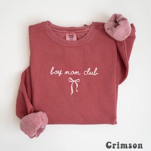 K&ouml;nnte beinhalten: Karmesinrotes Sweatshirt mit dem Text "boy mom club" in wei&szlig;er Schrift und einer Schleifengrafik. Das Sweatshirt hat einen Rundhalsausschnitt und ein weiches, flauschiges Futter an den Manschetten. Das Wort "Crimson" ist unten aufgedruckt.