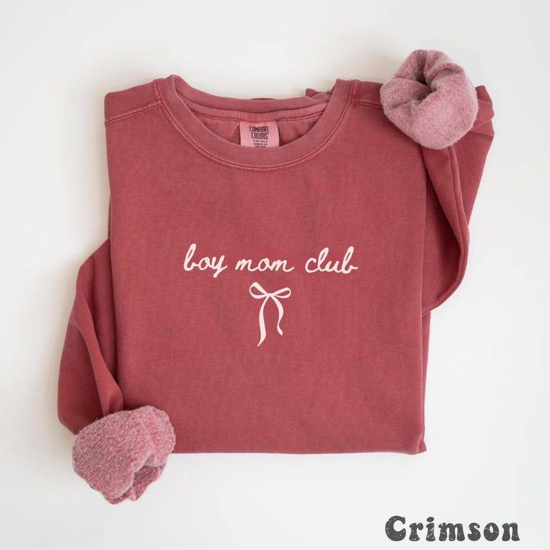 K&ouml;nnte beinhalten: Karmesinrotes Sweatshirt mit dem Text "boy mom club" in wei&szlig;er Schrift und einer Schleifengrafik. Das Sweatshirt hat einen Rundhalsausschnitt und ein weiches, flauschiges Futter an den Manschetten. Das Wort "Crimson" ist unten aufgedruckt.