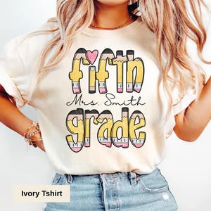 Puede incluir: Camiseta marfil con la frase "Fifth Grade Mrs. Smith" en un diseño de lápices. El texto es en amarillo, negro y rosa, con un pequeño corazón rosa encima de la palabra "Fifth". La camiseta es de material suave.