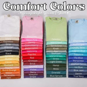 May include: Stacks of folded Comfort Colors heavyweight unisex t-shirts in various colors. The image displays a range of hues, including white, pink, green, and blue, with color names like "Blossom" and "Chambray" listed.