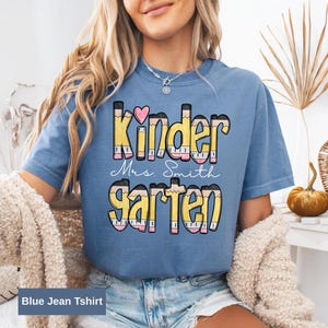 May include: A blue jean t-shirt with the words "Kindergarten Mrs. Smith" in a colorful pencil-themed design. The word "Kinder" has a pink heart above the letter "i". The shirt is a light blue color.