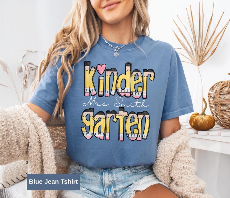 May include: A blue jean t-shirt with the words "Kindergarten Mrs. Smith" in a colorful pencil-themed design. The word "Kinder" has a pink heart above the letter "i". The shirt is a light blue color.
