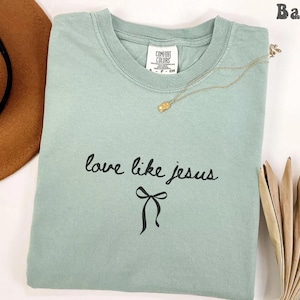May include: A light green t-shirt with the words "love like jesus" in black script and a black bow graphic. A gold necklace with a small pendant rests on the shirt. A brown hat is partially visible in the upper left corner.