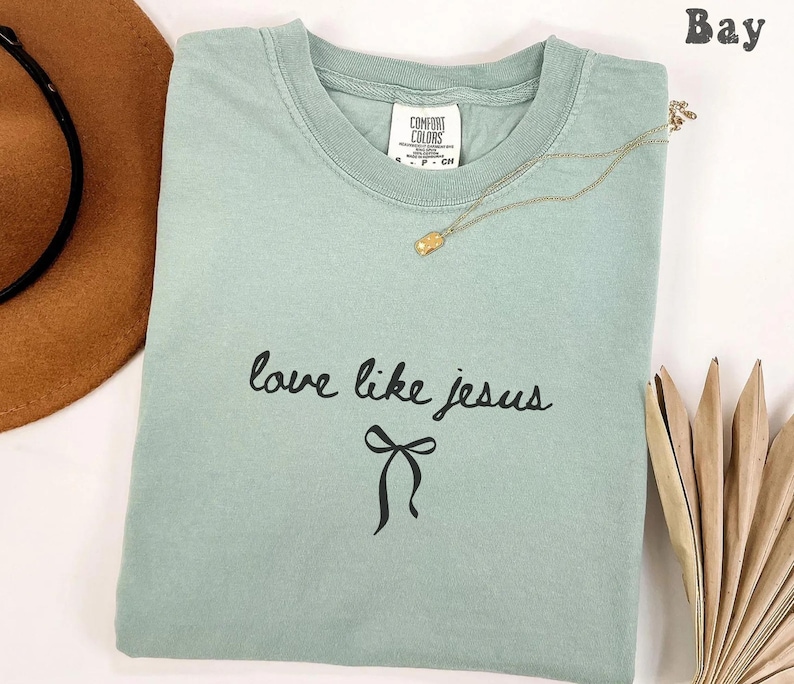 May include: A light green t-shirt with the words "love like jesus" in black script and a black bow graphic. A gold necklace with a small pendant rests on the shirt. A brown hat is partially visible in the upper left corner.