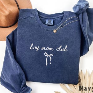 K&ouml;nnte beinhalten: Marineblaues Sweatshirt mit wei&szlig;em Schriftzug "boy mom club" und einer Schleifengrafik. Eine goldene Halskette liegt auf der Vorderseite. Das Sweatshirt ist aus weichem Material.
