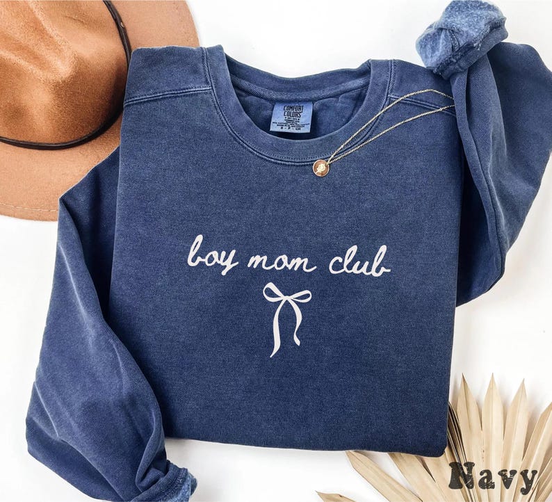K&ouml;nnte beinhalten: Marineblaues Sweatshirt mit wei&szlig;em Schriftzug "boy mom club" und einer Schleifengrafik. Eine goldene Halskette liegt auf der Vorderseite. Das Sweatshirt ist aus weichem Material.