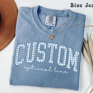 Puede incluir: Una camiseta color azul jean con cuello redondo y mangas cortas. El frente presenta la palabra CUSTOM en letras grandes de bloque con un patrón de cuadros vichy azul claro y blanco, seguido del texto optional line en cursiva blanca.