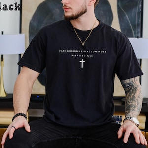 Puede incluir: Camiseta negra con el texto "FATHERHOOD IS KINGDOM WORK" y una cruz blanca. El texto "Proverbios 22:6" est&aacute; debajo. La camiseta es de color negro liso.