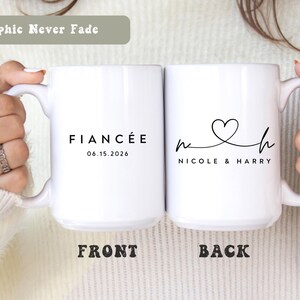 Puede incluir: Dos tazas de cerámica blancas. Una taza dice "FIANCÉE" con la fecha "06.15.2026". La otra taza tiene los nombres "NICOLE & HARRY" con un diseño de corazón. Las tazas son sostenidas por una persona.