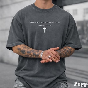 May include: Dark gray t-shirt with the text "FATHERHOOD IS KINGDOM WORK" and a white cross. The shirt is a relaxed fit with short sleeves. The text "Proverbs 22:6" is below the main text.