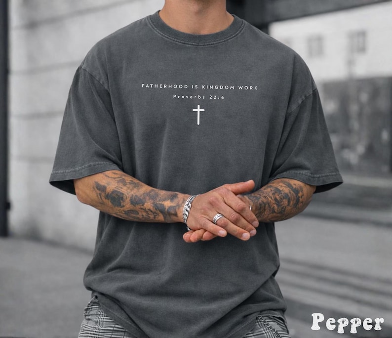 Puede incluir: Camiseta gris oscuro con el texto "FATHERHOOD IS KINGDOM WORK" y una cruz blanca. La camiseta es de corte holgado con mangas cortas. El texto "Proverbios 22:6" est&aacute; debajo.