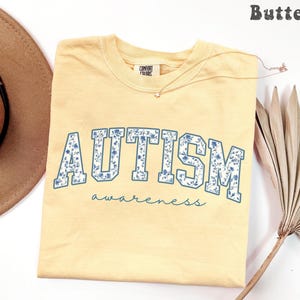 May include: A light yellow t-shirt with the word "AUTISM" in blue floral letters and the word "awareness" in a script font. The shirt is a Comfort Colors brand. A gold necklace with a small pendant is on the shirt.