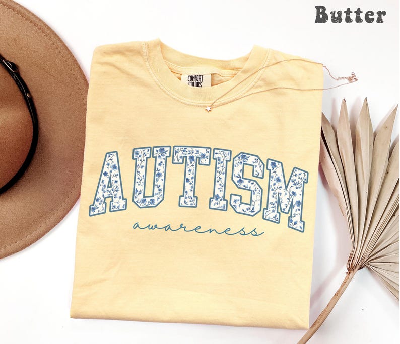 May include: A light yellow t-shirt with the word "AUTISM" in blue floral letters and the word "awareness" in a script font. The shirt is a Comfort Colors brand. A gold necklace with a small pendant is on the shirt.