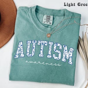 May include: Light green t-shirt with the word "AUTISM" in blue floral letters. The word "awareness" is written in cursive below. A silver necklace with a pendant is on the shirt.