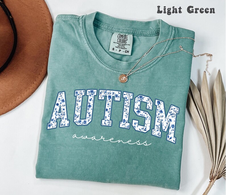May include: Light green t-shirt with the word "AUTISM" in blue floral letters. The word "awareness" is written in cursive below. A silver necklace with a pendant is on the shirt.