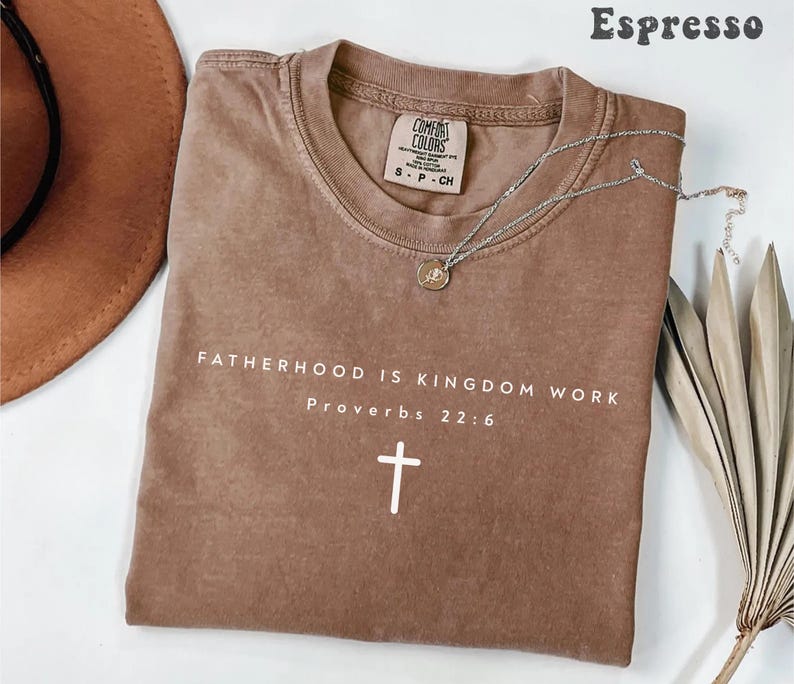 Puede incluir: Camiseta color espresso con el texto "FATHERHOOD IS KINGDOM WORK Proverbs 22:6" y una cruz blanca. Un collar plateado con un colgante descansa sobre la camiseta. La camiseta est&aacute; doblada y tiene una etiqueta Comfort Colors.