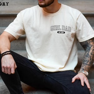 May include: Ivory-colored t-shirt with the text "GIRL DAD EST. 2025" in black. The shirt is a short-sleeved crew neck style. The model is wearing black jeans and a watch.