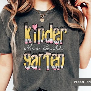 May include: Dark gray t-shirt with the words "Kinder Mrs. Smith Garten" in a colorful pencil design. The word "Kinder" has a pink heart above it. The shirt is a comfortable, casual style.