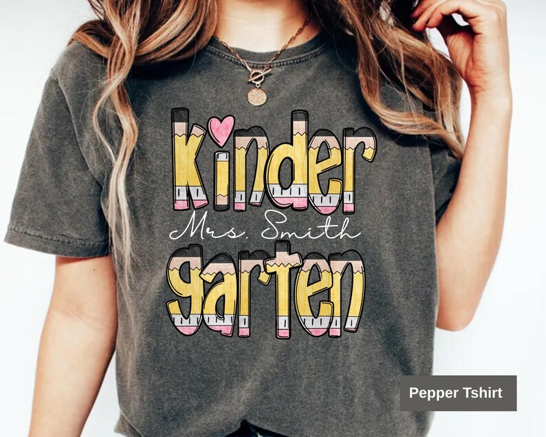 May include: Dark gray t-shirt with the words "Kinder Mrs. Smith Garten" in a colorful pencil design. The word "Kinder" has a pink heart above it. The shirt is a comfortable, casual style.