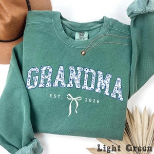 May include: A light green sweatshirt with the word "GRANDMA" in white letters filled with a blue floral pattern. Below the word "GRANDMA" is the text "EST. 2026" and a white bow graphic. A gold necklace is draped across the sweatshirt.