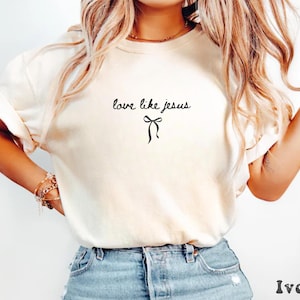 May include: Ivory-colored t-shirt with the phrase "love like jesus" in a handwritten font, accompanied by a simple bow graphic. The shirt has short sleeves and a relaxed fit. The bottom right corner of the shirt has the word "Ivory".