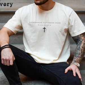 Puede incluir: Camiseta de color marfil con el texto "FATHERHOOD IS KINGDOM WORK" y un s&iacute;mbolo de cruz. El texto "Proverbios 22:6" est&aacute; debajo. La camiseta es de cuello redondo y tiene mangas cortas.
