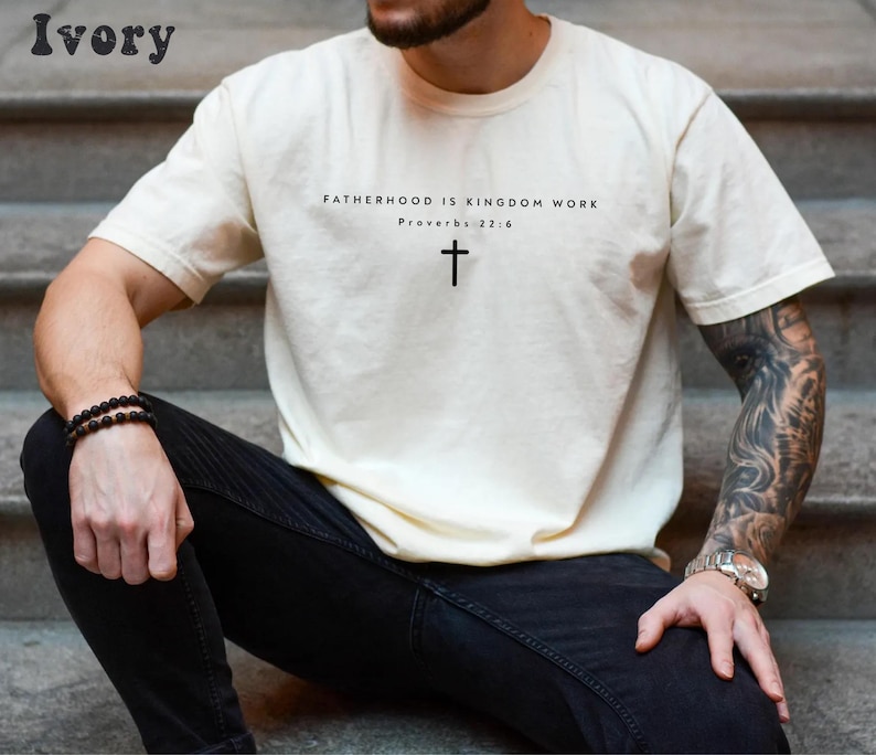 Puede incluir: Camiseta de color marfil con el texto "FATHERHOOD IS KINGDOM WORK" y un s&iacute;mbolo de cruz. El texto "Proverbios 22:6" est&aacute; debajo. La camiseta es de cuello redondo y tiene mangas cortas.