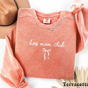 K&ouml;nnte beinhalten: Terracottafarbenes Sweatshirt mit den Worten "boy mom club" in wei&szlig;er Schrift und einer Schleifengrafik. Das Sweatshirt hat einen Rundhalsausschnitt und lange &Auml;rmel. Eine goldene Halskette ist &uuml;ber die Vorderseite drapiert. Ein brauner Hut ist im Hintergrund.
