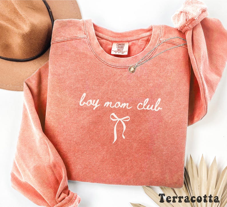K&ouml;nnte beinhalten: Terracottafarbenes Sweatshirt mit den Worten "boy mom club" in wei&szlig;er Schrift und einer Schleifengrafik. Das Sweatshirt hat einen Rundhalsausschnitt und lange &Auml;rmel. Eine goldene Halskette ist &uuml;ber die Vorderseite drapiert. Ein brauner Hut ist im Hintergrund.