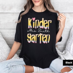 May include: Black t-shirt with the words "Kinder Mrs. Smith Garten" in a colorful pencil-themed design. A pink heart is above the word "Kinder". The shirt is a crew neck style.