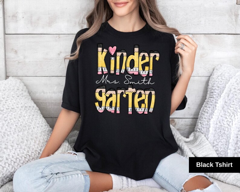 May include: Black t-shirt with the words "Kinder Mrs. Smith Garten" in a colorful pencil-themed design. A pink heart is above the word "Kinder". The shirt is a crew neck style.