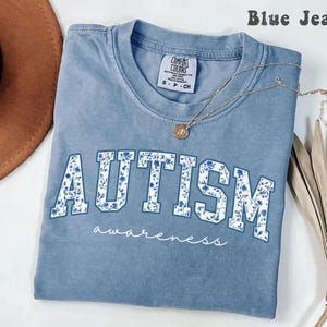 Blue Jean Comfort Colors shirt with blue floral patterned “AUTISM” text and awareness script underneath, designed for autism support, neurodiversity advocacy, and everyday casual wear.